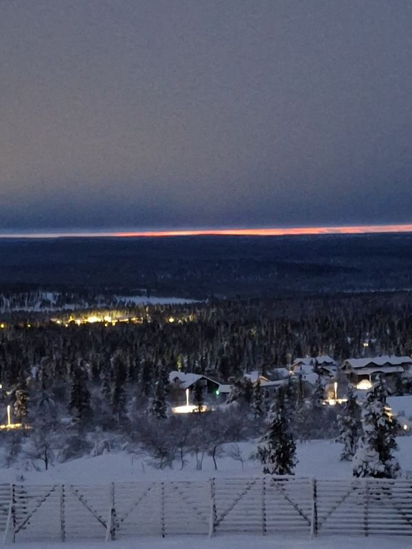 Schneelandschaft mit Himmel, Ort und Wald . Der Himmel gelblichgrau. Über dem Horizont ein zarter orangener Streifen und darüber ein dunkelblauer Bereich.