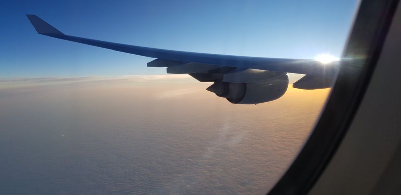 Blick aus dem Flugzeugfenster auf die Wolkendecke und den Tragflügel, hinter dem die Sonne hervorlugt